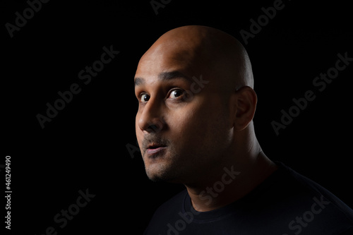 Portrait of a bald man with uncanny expression gazing elsewhere against black background.