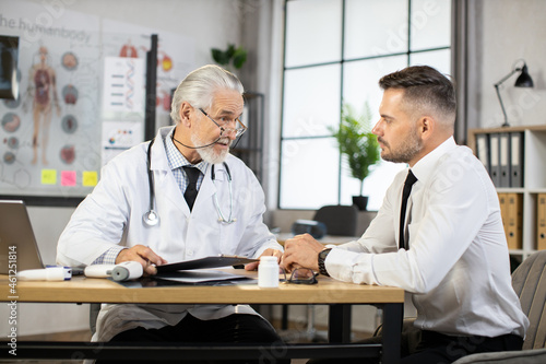Caucasian businessman in formal clothes and senior male doctor in lab coat sitting at medical cabinet and talking. Patient receiving professional consultation and check up a modem clinic.
