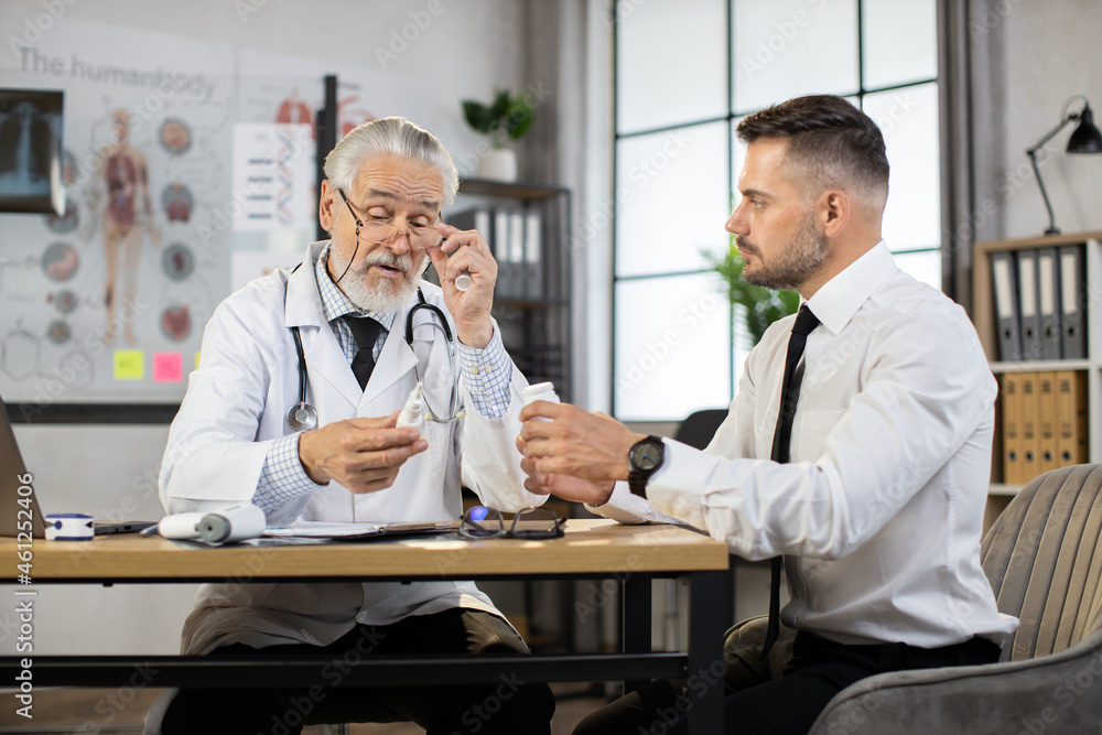 Competent senior doctor holding various pills in hands and explaining prescription to male patient while sitting at medical cabinet. Professional consultation at modern clinic. Competent senior doctor holding various pills in hands and explaining prescription to male patient while sitting at medical cabinet. Professional consultation at modern clinic.