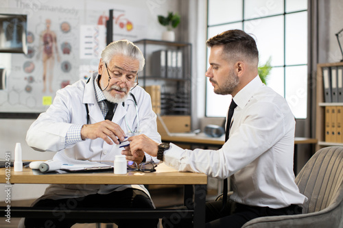 Caucasian aged doctor checking saturation with fingertip pulse oximeter of male patient in formal clothes. Male therapist examining businessman during appointment at modern clinic.