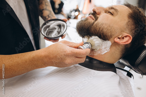 Close-up male hands with shaving brush prepare beard of customer for cutting at barbershop. Beauty, selfcare, style, fashion and male cosmetics concept.