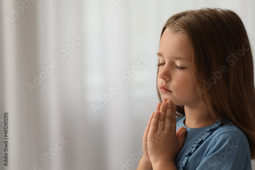 Cute little girl with hands clasped together praying indoors. Space for text