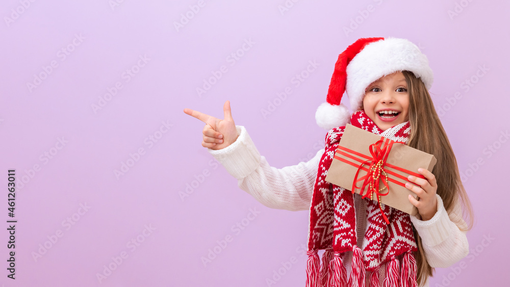 Obraz premium A beautiful little girl is very happy about Christmas. A child in a New Year's hat and with a gift in his hands points his finger to the side.