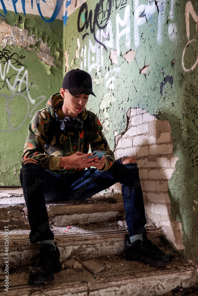 Teenager with black cap sitting on stairs in ruined building and using smarthphone