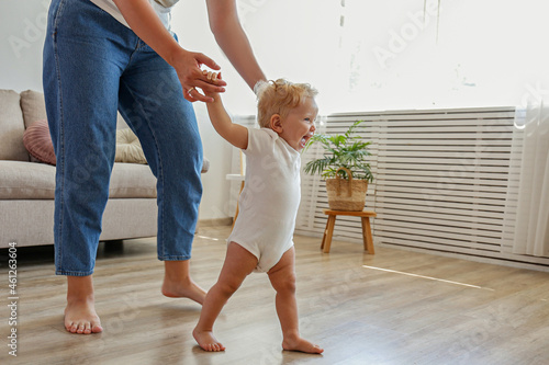 First steps of a little girl. A blond toddler learning to walk at home with the help of loving mother. Close up, copy space for text, background.