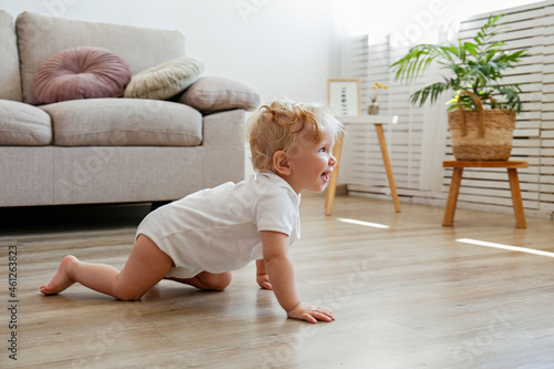 One year old child learning to crawl on the wooden floor of living room. Interior background. Adorable blonde little girl in pink crawlers at home. Close up, copy space for text. photo