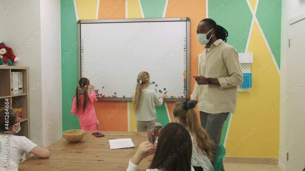 African American teacher in a mask teaches a group of masked children learning English. School for Children, Teaching Adolescents, Gain Knowledge, Learn the Language. African American teacher in a mask teaches a group of masked children learning English. School for Children, Teaching Adolescents, Gain Knowledge, Learn the Language.