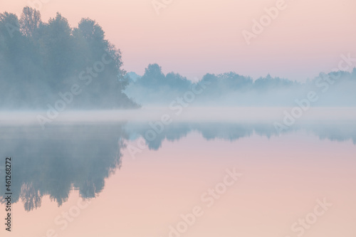 Dawn over the foggy lake. Beautiful dreamy view. Pink sky just before the sunrise and fog over water and trees with reflections on the river bank.