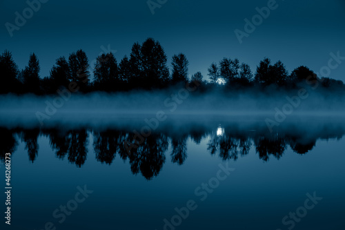 Night mystical scenery. Full moon through the tree branches, rising over the foggy river and its reflection in the still water. © stone36