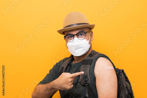 Vaccinated. Traveler asian man wearing mask showing arm after coronavirus Vaccination isolated yellow Background. Backpacker Indian man getting a vaccination covid-19 immunization