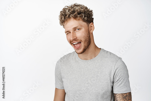 Handsome macho man with blond curly hair and bristle, winking at camera and smiling, standing in gray t-shirt against white background