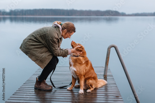 Handsome caucasian girl caress brown Nova Scotia Duck Tolling Retriever. Woman with pet sitting on pier on lake and look into each other eyes. Female travelling with dog. Selective focus. Dog loyalty.
