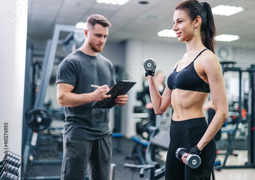 Instructor male writing in clipboard while young woman exercising with dumbbells at gym. Personal trainer checks the results of a slim female in the sport centre
