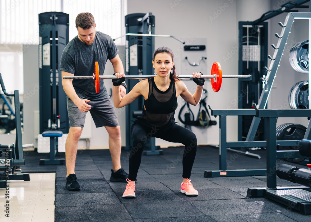 Sport, bodybuilding, lifestyle and people concept. Young man and woman with barbell flexing muscles and making shoulder press squat in gym. Trainer and fitness girl are having workout at gym Sport, bodybuilding, lifestyle and people concept. Young man and woman with barbell flexing muscles and making shoulder press squat in gym. Trainer and fitness girl are having workout at gym