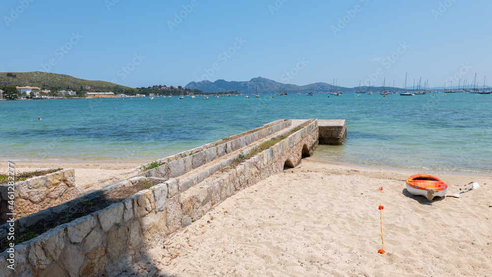 A beautiful beach in the bay of Pollenca on the Mediterranean island of Mallorca. Many boats are tied to buoys in front of the beach. Mountains can be seen in the background.