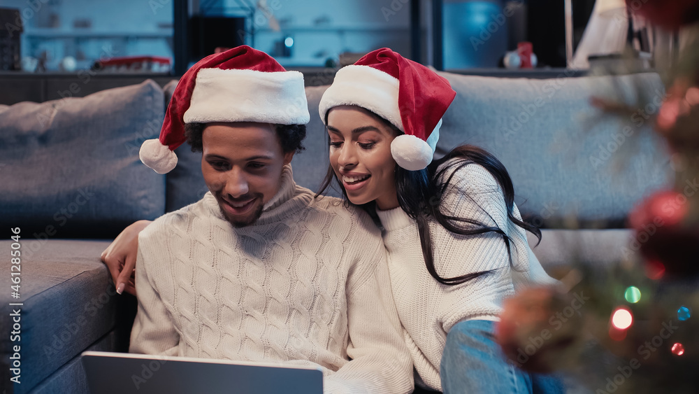 cheerful african american couple in santa hats using laptop near blurred christmas tree