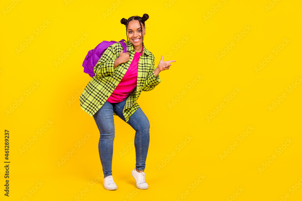 Full length photo of joyful positive afro american woman wear bag point finger empty space isolated on yellow color background Full length photo of joyful positive afro american woman wear bag point finger empty space isolated on yellow color background