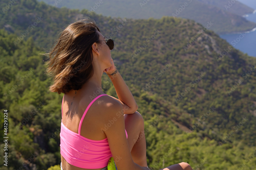 A young woman in a bright pink top sits on the top of a mountain with a beautiful view of the mountains and the mediterranean sea in southern Turkey