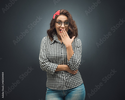 Surprised girl looks to camera over dark gray - black background. Cheerful young woman covering her mouth with hand.