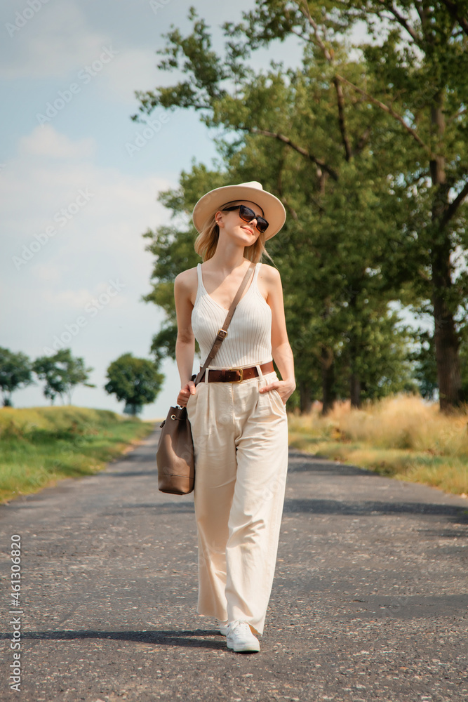 Stylish woman in hat, sunglasses and white clothes walking on a rural road