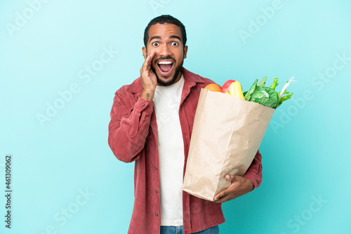Young latin man holding a grocery shopping bag isolated on blue background with surprise and shocked facial expression