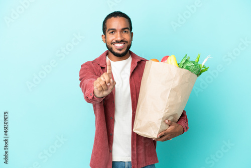 Young latin man holding a grocery shopping bag isolated on blue background showing and lifting a finger