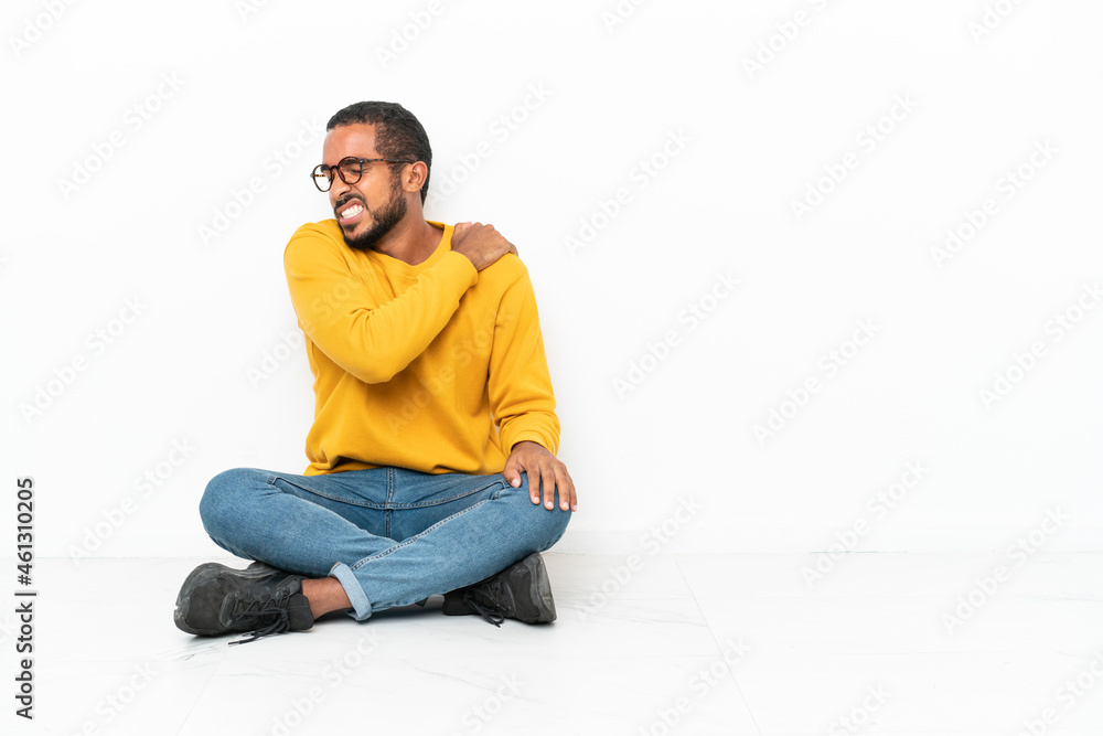 Young Ecuadorian man sitting on the floor isolated on white wall suffering from pain in shoulder for having made an effort