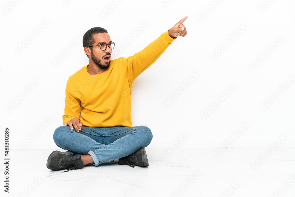 Young Ecuadorian man sitting on the floor isolated on white wall pointing away