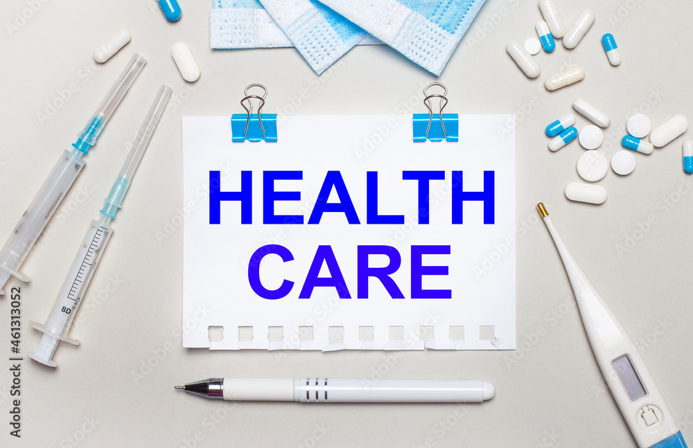 Fototapeta premium On a light gray background, blue medical masks, syringes, an electronic thermometer, pills, a pen and a notebook with the inscription HEALTH CARE. Medical concept
