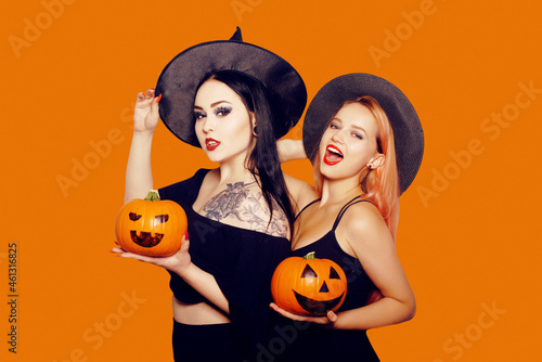 Two girls witches in hats are holding pumpkins in their hands and smiling for the camera, posing, dancing. Women celebrate Halloween, photo on a red background.