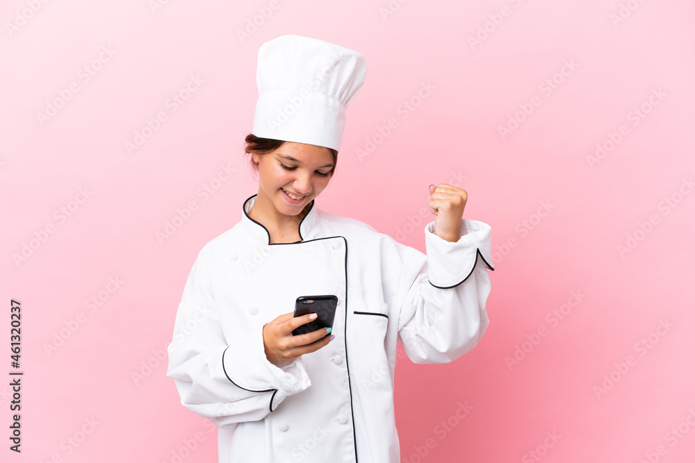 Little caucasian chef girl isolated on pink background with phone in victory position Little caucasian chef girl isolated on pink background with phone in victory position