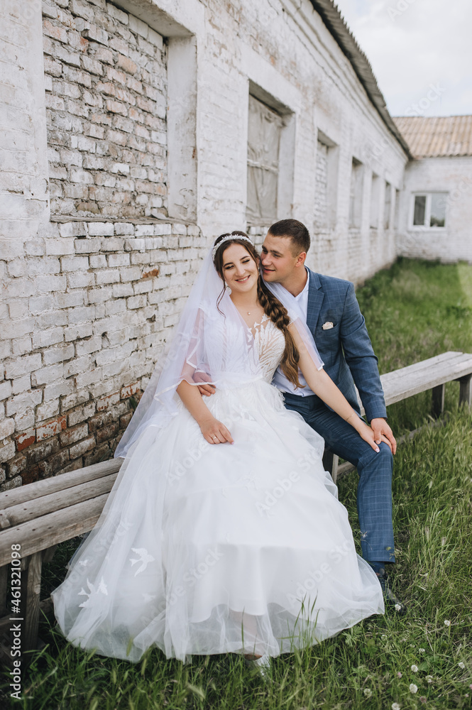 Stylish groom in a blue suit and a brunette bride in a white lace dress are sitting on a wooden bench in the village against the backdrop of a brick building, farm. Wedding photography. Stylish groom in a blue suit and a brunette bride in a white lace dress are sitting on a wooden bench in the village against the backdrop of a brick building, farm. Wedding photography.