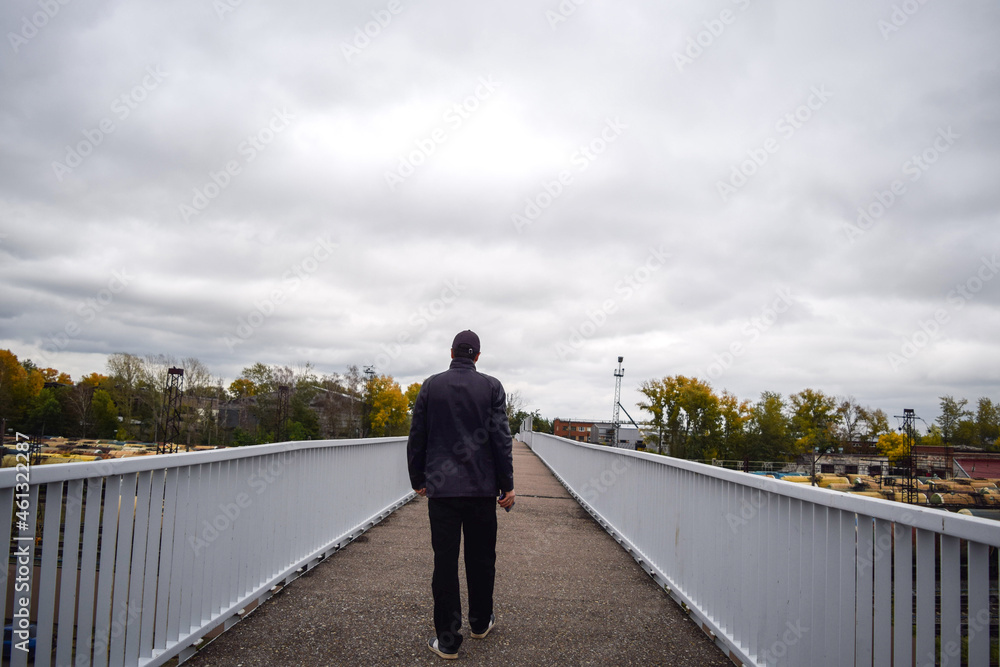 A man in a leather jacket and a cap walks across the bridge over the railway on an autumn evening