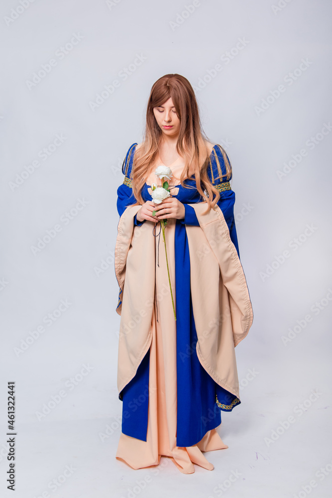 Fototapeta premium An attractive woman with long hair in a medieval, fantasy, blue and beige dress with long, large sleeves, posing with white roses in her hands, isolated on a white background.
