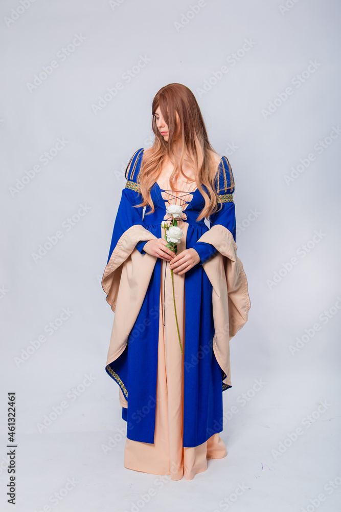 Fototapeta premium An attractive woman with long hair in a medieval, fantasy, blue and beige dress with long, large sleeves, posing with white roses in her hands, isolated on a white background.