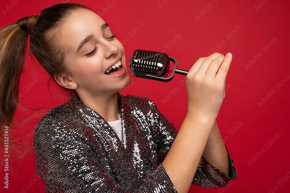 Photo shot of attractive happy positive brunette little girl wearing fashionable shine dress standing isolated over red background wall singing the song into the silver microphone looking to the side Photo shot of attractive happy positive brunette little girl wearing fashionable shine dress standing isolated over red background wall singing the song into the silver microphone looking to the side
