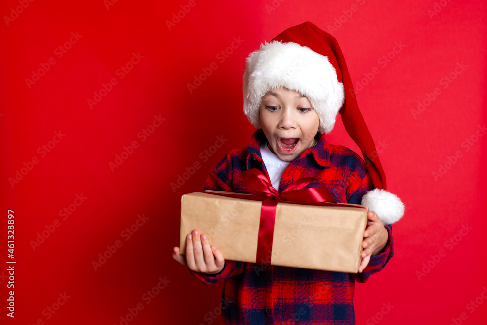 Happy holiday and Merry Christmas. Portrait of a funny surprised boy in a cap with gifts in his hands on a red background. A place for text. Happy holiday and Merry Christmas. Portrait of a funny surprised boy in a cap with gifts in his hands on a red background. A place for text.
