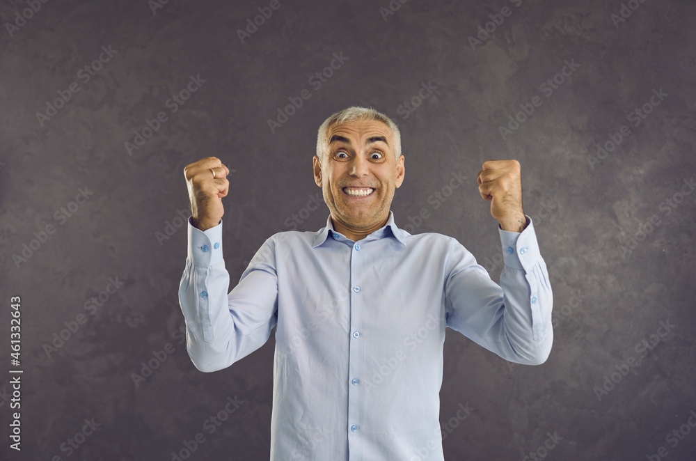 Portrait of happy emotional senior man who laughs madly and rejoices celebrating victory. Excited and joyful man with funny face clenching his fists in happiness standing on gray background. Portrait of happy emotional senior man who laughs madly and rejoices celebrating victory. Excited and joyful man with funny face clenching his fists in happiness standing on gray background.