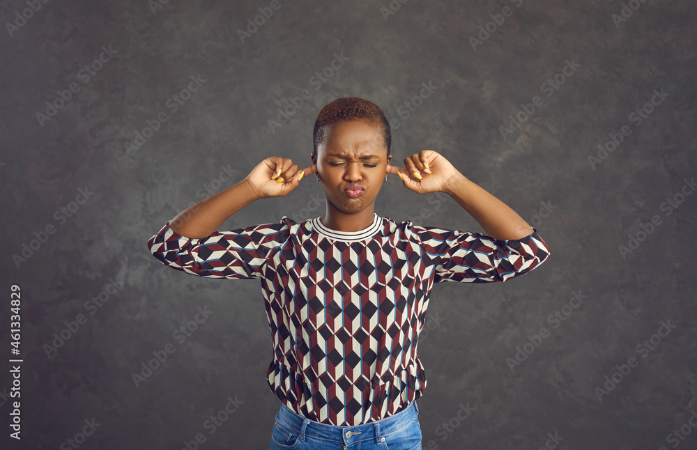 Capricious young African American woman covers her ears with her fingers so as not to hear anything. Woman with short hair dressed in jeans and a sweater stands with a frown on a gray background. - obrazy, fototapety, plakaty 