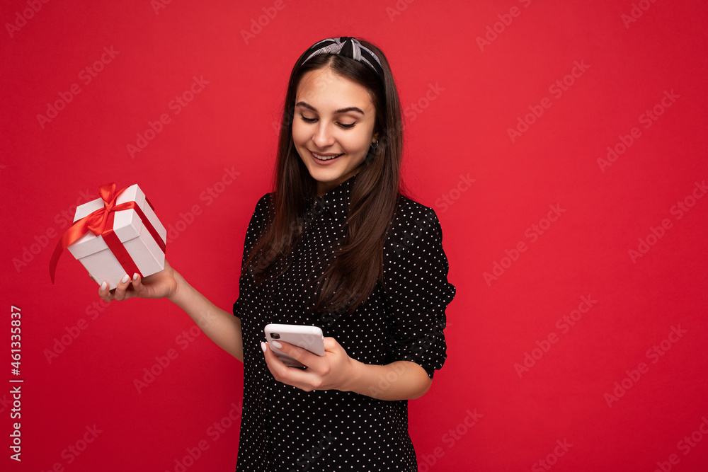 Photo shot of attractive cute positive smiling brunette girl isolated over red background wall wearing black blouse holding white gift box with red ribbon and looking at mobile phone screen display Photo shot of attractive cute positive smiling brunette girl isolated over red background wall wearing black blouse holding white gift box with red ribbon and looking at mobile phone screen display