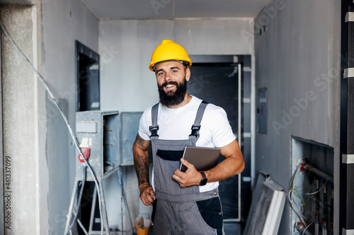 A happy construction worker with a helmet is standing in an unfinished building and holding a tablet under the armpit. He is checking on works on site. photo