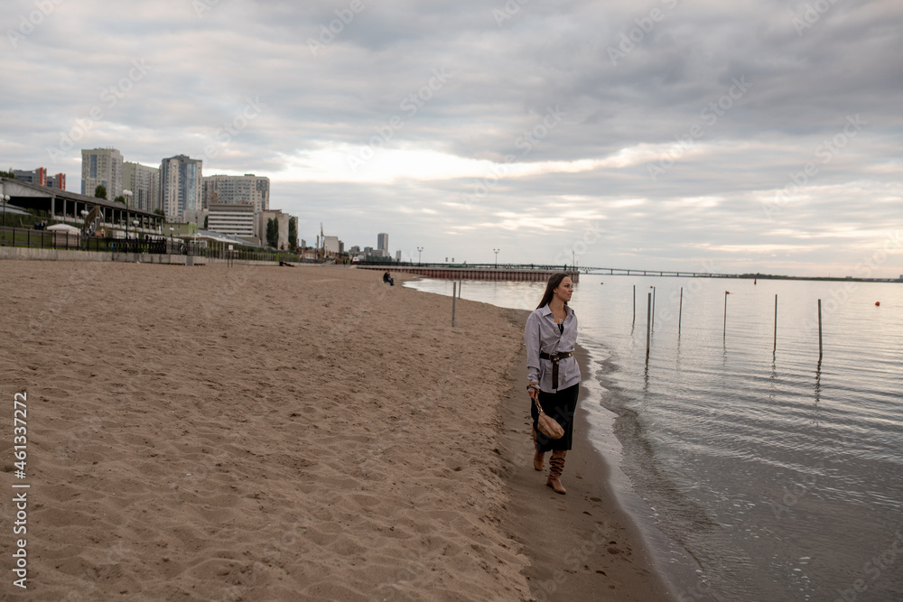 A beautiful young brunette woman walks on the beach in cool weather in a fashionable outfit