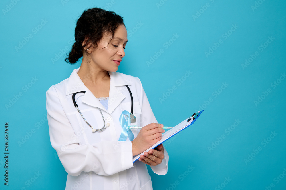 Middle aged mixed race woman, female doctor wearing a blue satin ribbon, symbolic bow color raising awareness in diabetes day , makingtes on a clipboard, isolated over blue background with copy space Middle aged mixed race woman, female doctor wearing a blue satin ribbon, symbolic bow color raising awareness in diabetes day , makingtes on a clipboard, isolated over blue background with copy space
