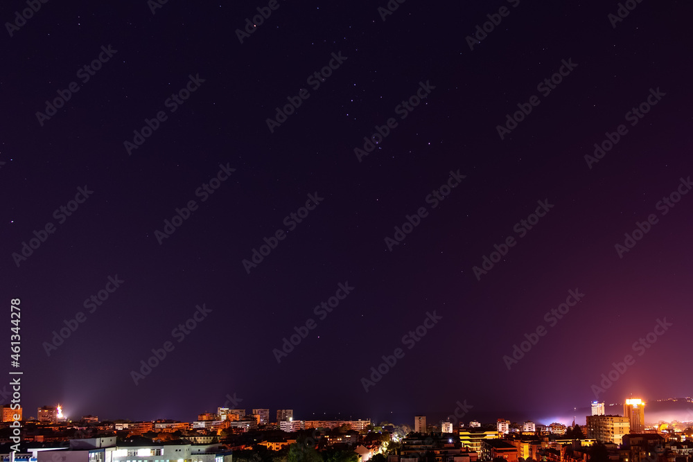 The constellation Orion shines brightly in the dark sky over illuminated city skyline at midnight. Clear starry sky over Varna in autumn. Night sky with bright stars over tranquil urban scene.