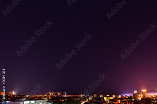 The constellation Orion shines brightly in the dark sky over illuminated city skyline at midnight. Clear starry sky over Varna in autumn. Night sky with bright stars over tranquil urban scene.