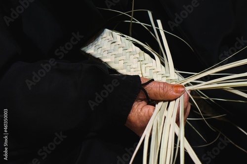 Hands of elderly arab woman artisan in traditional black dress close up weaving manually basket from dried organic palm leaves. Arabian traditional crafts. 