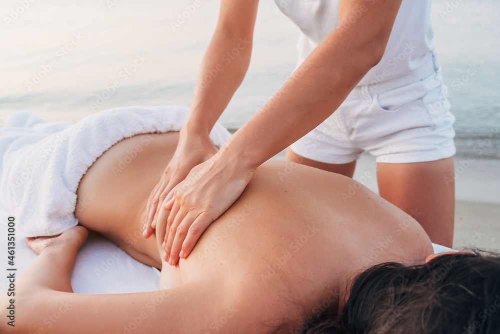 A girl massage therapist makes a back massage to a woman on the seashore. A woman lies on a massage table and is covered with a white towel and relaxes A girl massage therapist makes a back massage to a woman on the seashore. A woman lies on a massage table and is covered with a white towel and relaxes