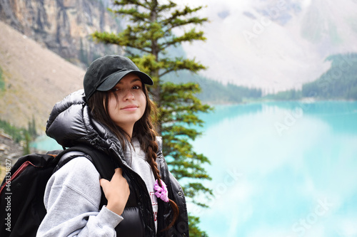 Young girl with backpack standing in front of turquoise lake and mountains smiling looking at camera © Julia