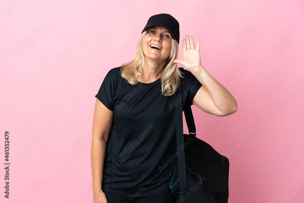 Middle age woman with sport bag isolated on pink background listening to something by putting hand on the ear