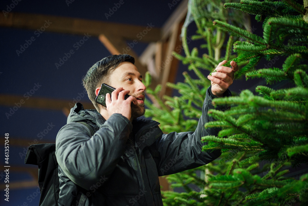 Festive shopping, selection and purchase of New Years attributes. Caucasian man speaks on phone and advises on choosing Christmas tree on evening of New Year Eve. Male buys spruce and uses smartphone. Festive shopping, selection and purchase of New Years attributes. Caucasian man speaks on phone and advises on choosing Christmas tree on evening of New Year Eve. Male buys spruce and uses smartphone.